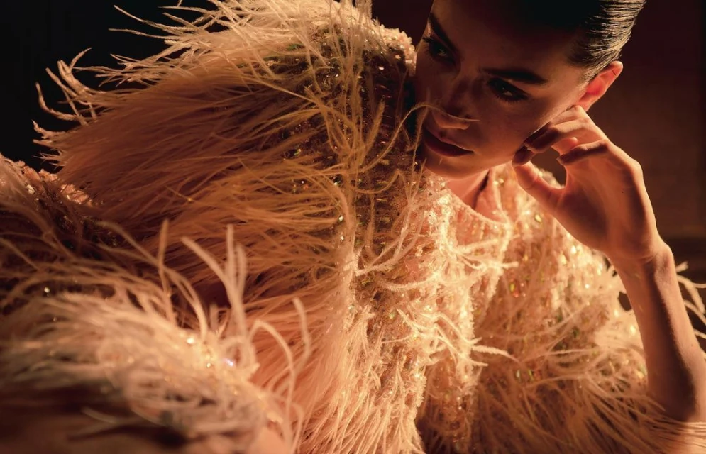 Elegant woman with hand to face, wearing feathered couture in warm, moody light, soft focus, beauty editorial. Glamour aesthetic.