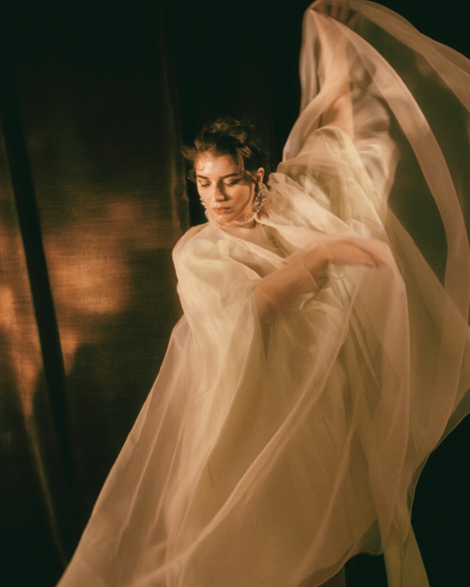 Woman in sheer white flowing fabric dancing with arms raised against a dark background, photographed by Noemi Ottilia Szabo in Paris.