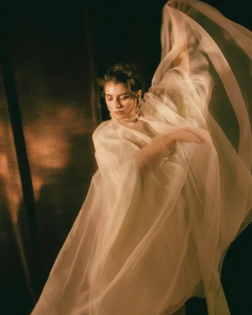Woman in sheer white flowing fabric dancing with arms raised against a dark background, photographed by Noemi Ottilia Szabo in Paris.