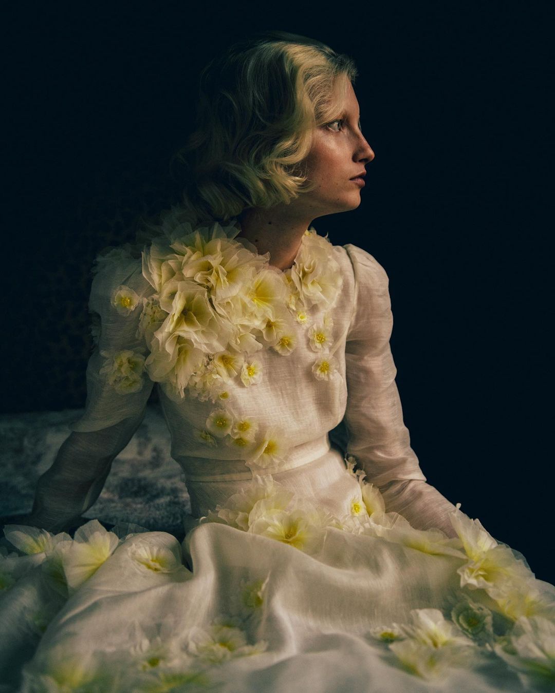 Young woman in a white couture gown with floral embellishments poses in a dark studio, side profile, soft lighting, for a fashion editorial.