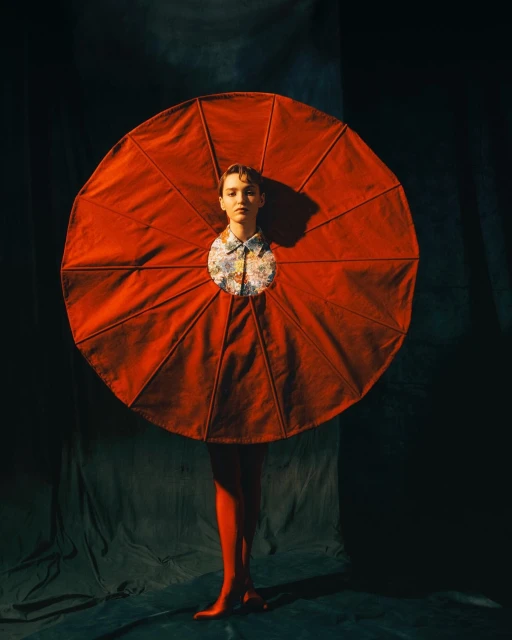 Woman in red textile art piece, wearing a floral blouse and red tights, poses against a dark backdrop in a portrait.
