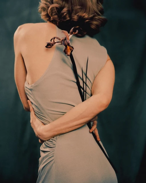 A woman with arms crossed behind her back wearing a light-grey dress and an iris flower in the photo studio.