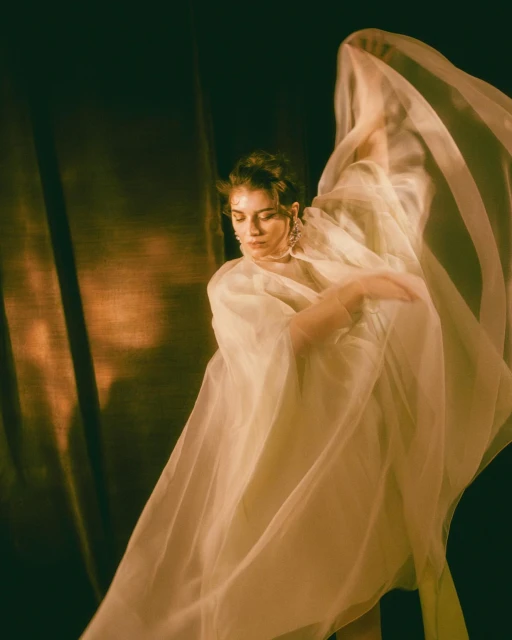 Woman in sheer white dress with tulle, posing in front of a textured brown background, bathed in soft, golden-toned light.