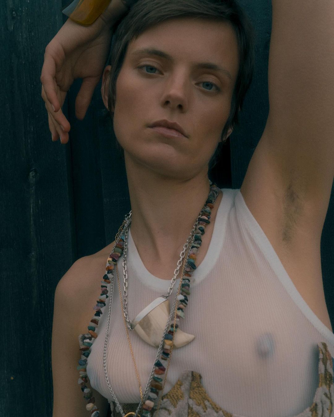 Portrait of a woman with short hair, wearing a white tank top and stone necklaces, posing against a dark green wooden wall outdoors.