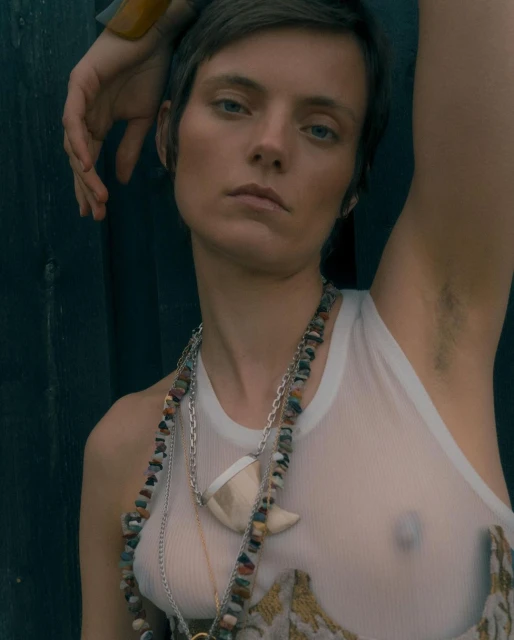 Portrait of a woman with short hair, wearing a white tank top and stone necklaces, posing against a dark green wooden wall outdoors.