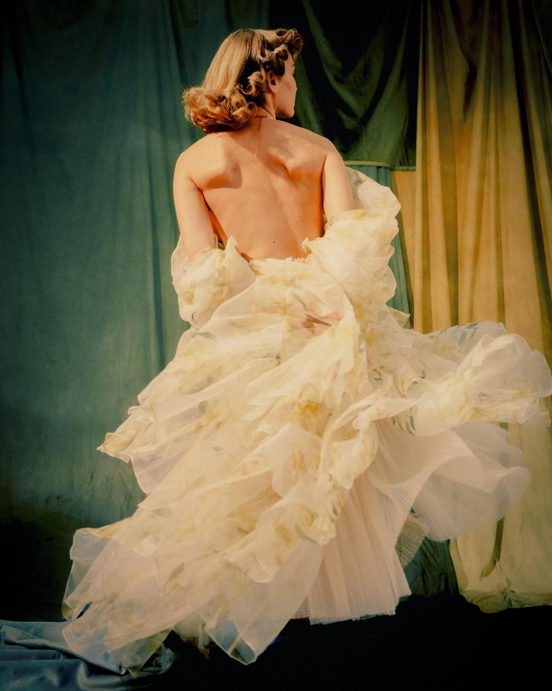 A woman in a flowing gown is seen from the back, posing against a draped green and tan background in a fashion or beauty shot.