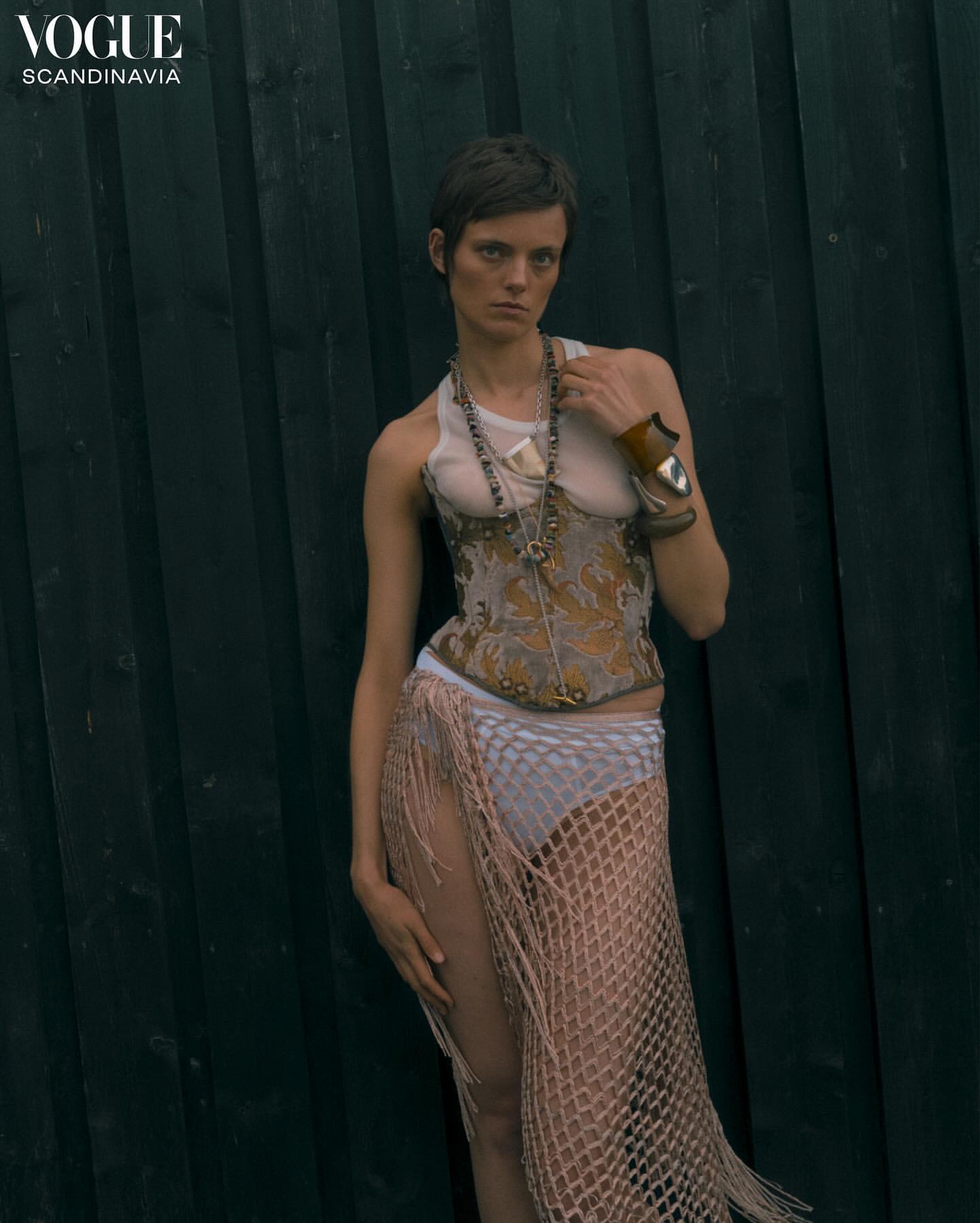 Model with short dark hair wearing a patterned corset and fringed wrap skirt against a dark wooden slat wall