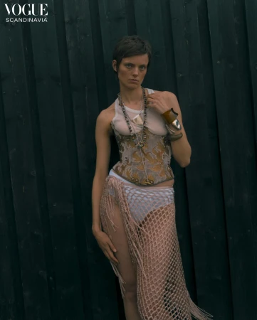Model with short dark hair wearing a patterned corset and fringed wrap skirt against a dark wooden slat wall
