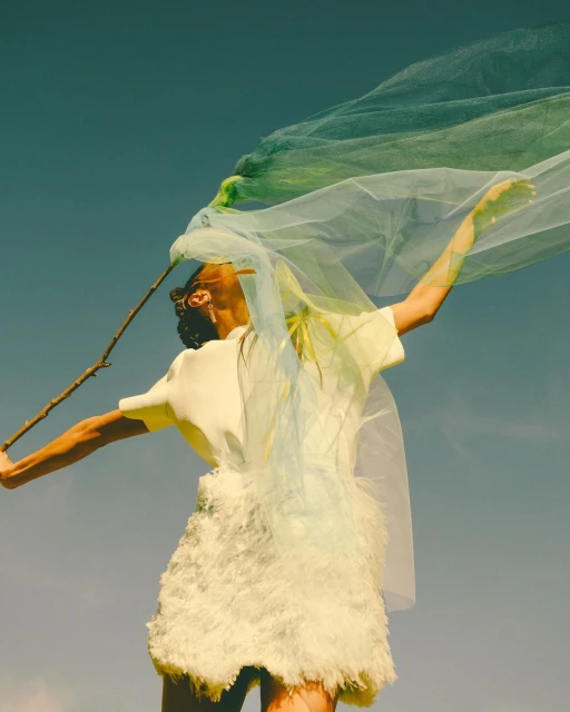 Woman in a cream dress and green tulle stands against a blue sky, holding a branch, creating a surreal and artistic fashion portrait.