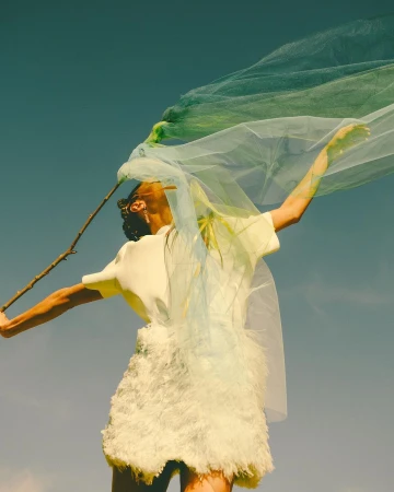 Woman poses in white dress with tulle against blue sky, holding stick. Fashion-editorial shot, light, airy and ethereal aesthetic.