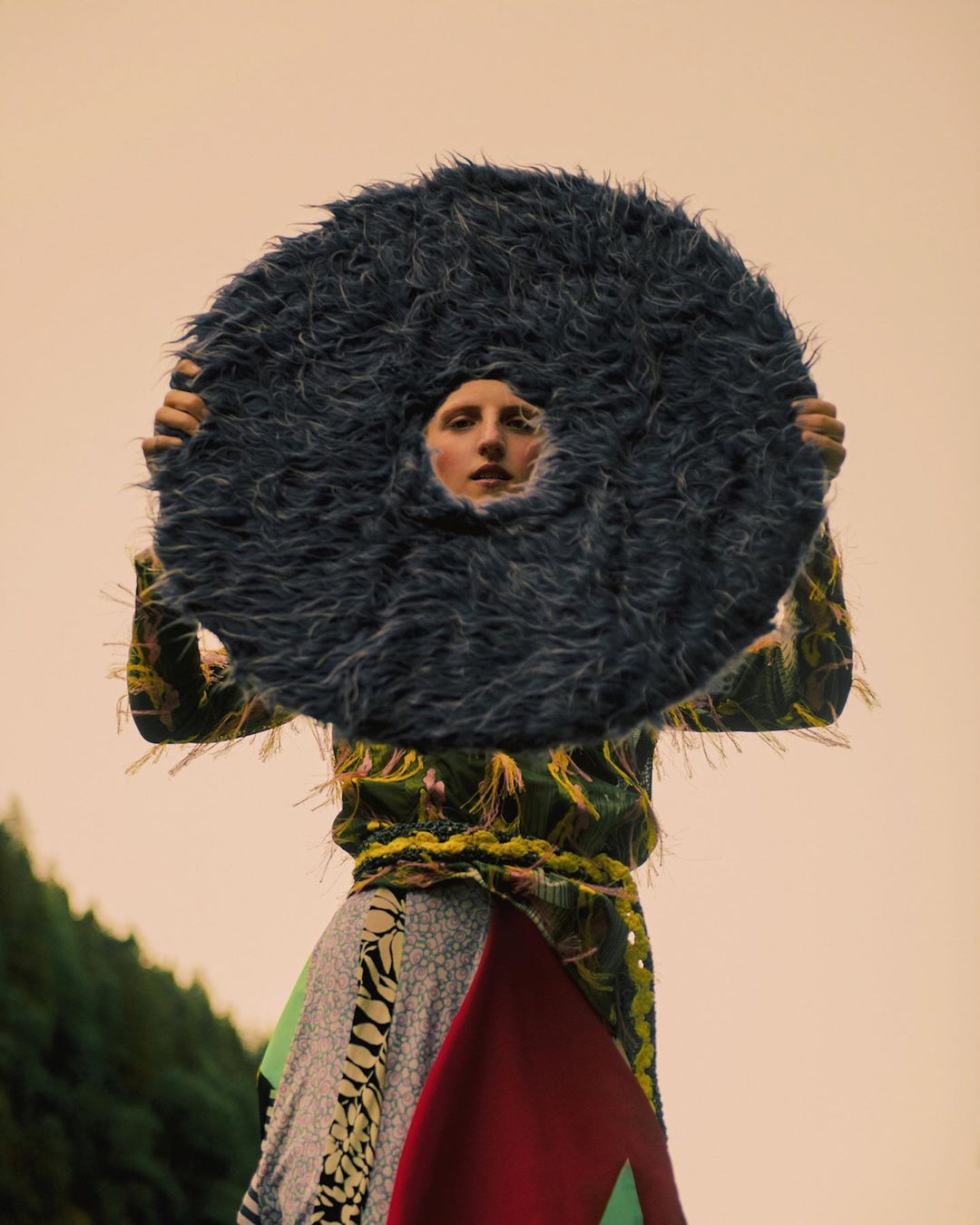 Model posing with a large, round, furry prop in an outdoor setting; the person wears colorful clothing and stands against a peach-toned sky.