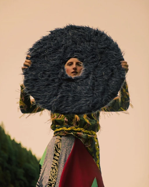 Model posing with a large, round, furry prop in an outdoor setting; the person wears colorful clothing and stands against a peach-toned sky.
