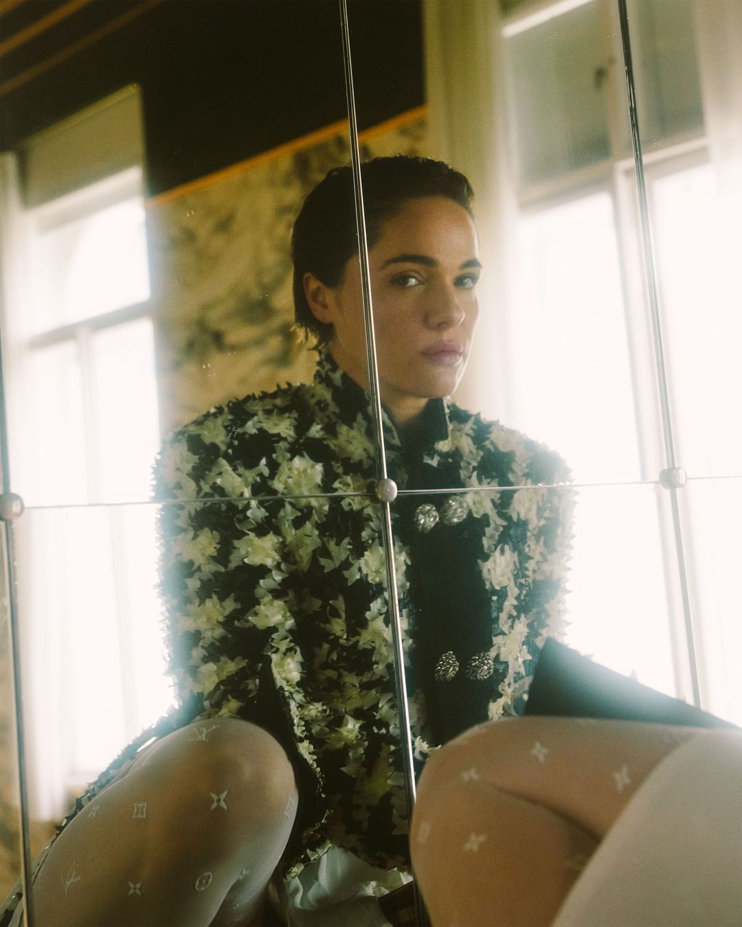 A woman with short dark hair poses indoors, wearing a floral jacket, hosiery with logos, and standing behind a glass barrier or mirror.