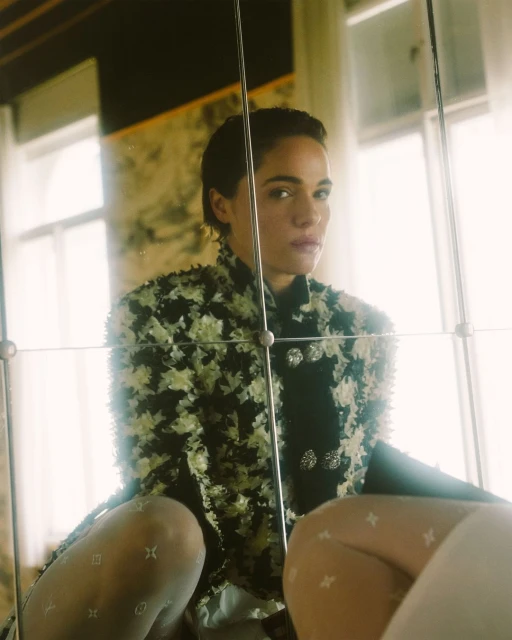 A woman with short dark hair poses indoors, wearing a floral jacket, hosiery with logos, and standing behind a glass barrier or mirror.
