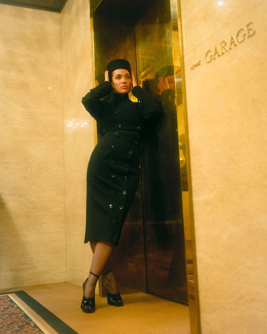 Stylish adult female in black hat and dress posing near golden elevator with 'Garage' sign, wearing black patent leather shoes.