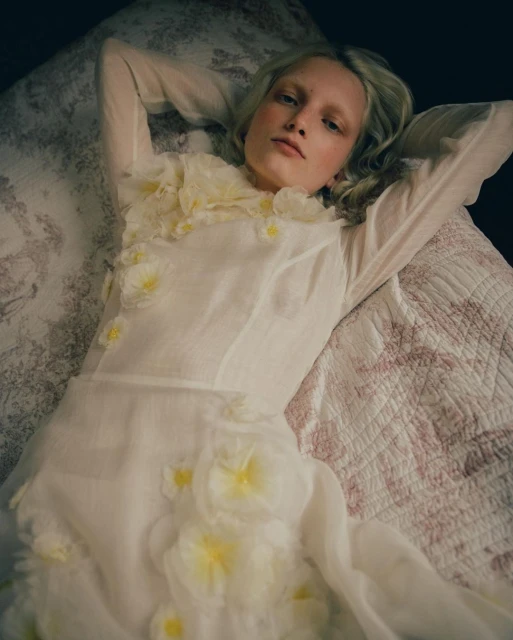 Woman in a white dress with flower appliques lays on a floral quilt, looking at the camera in a bright, indoor setting.