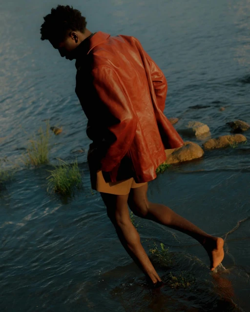 A dark-skinned man in a red jacket wading through water with rocks and foliage at the edge, photographed in a natural setting.
