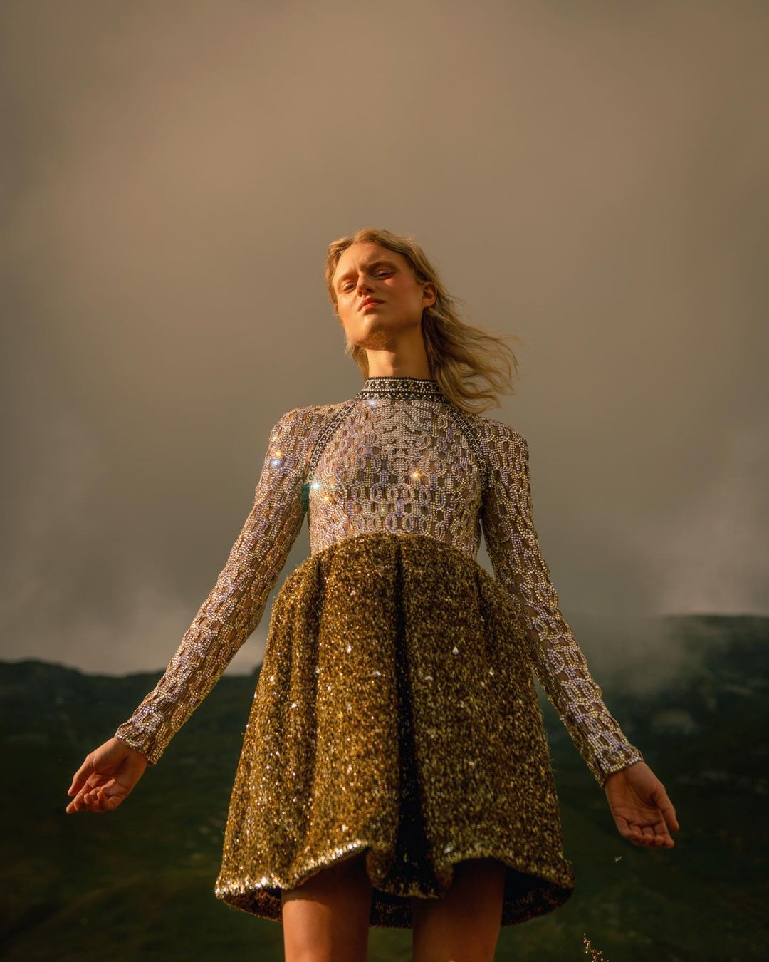 Young woman in elegant sequin dress standing in outdoor landscape, wind blowing hair. Low angle, golden hour, dramatic sky.