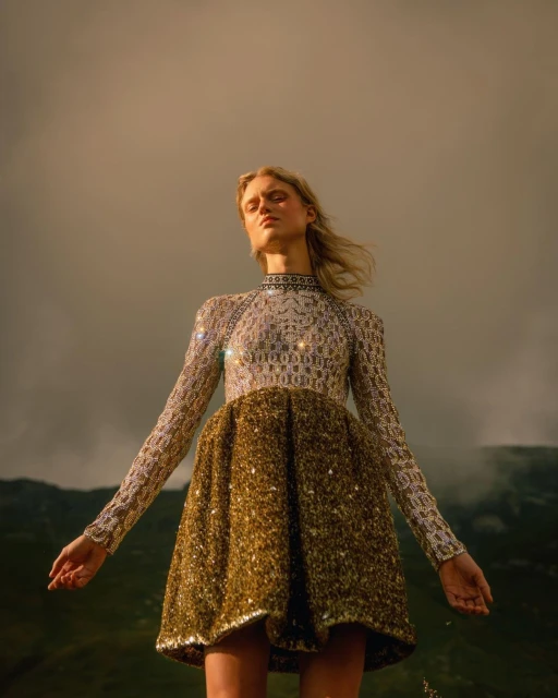 Young woman in elegant sequin dress standing in outdoor landscape, wind blowing hair. Low angle, golden hour, dramatic sky.