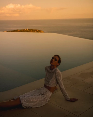 Woman in a shimmering two-piece outfit poses by an infinity pool with ocean and island views during golden hour with soft lighting.