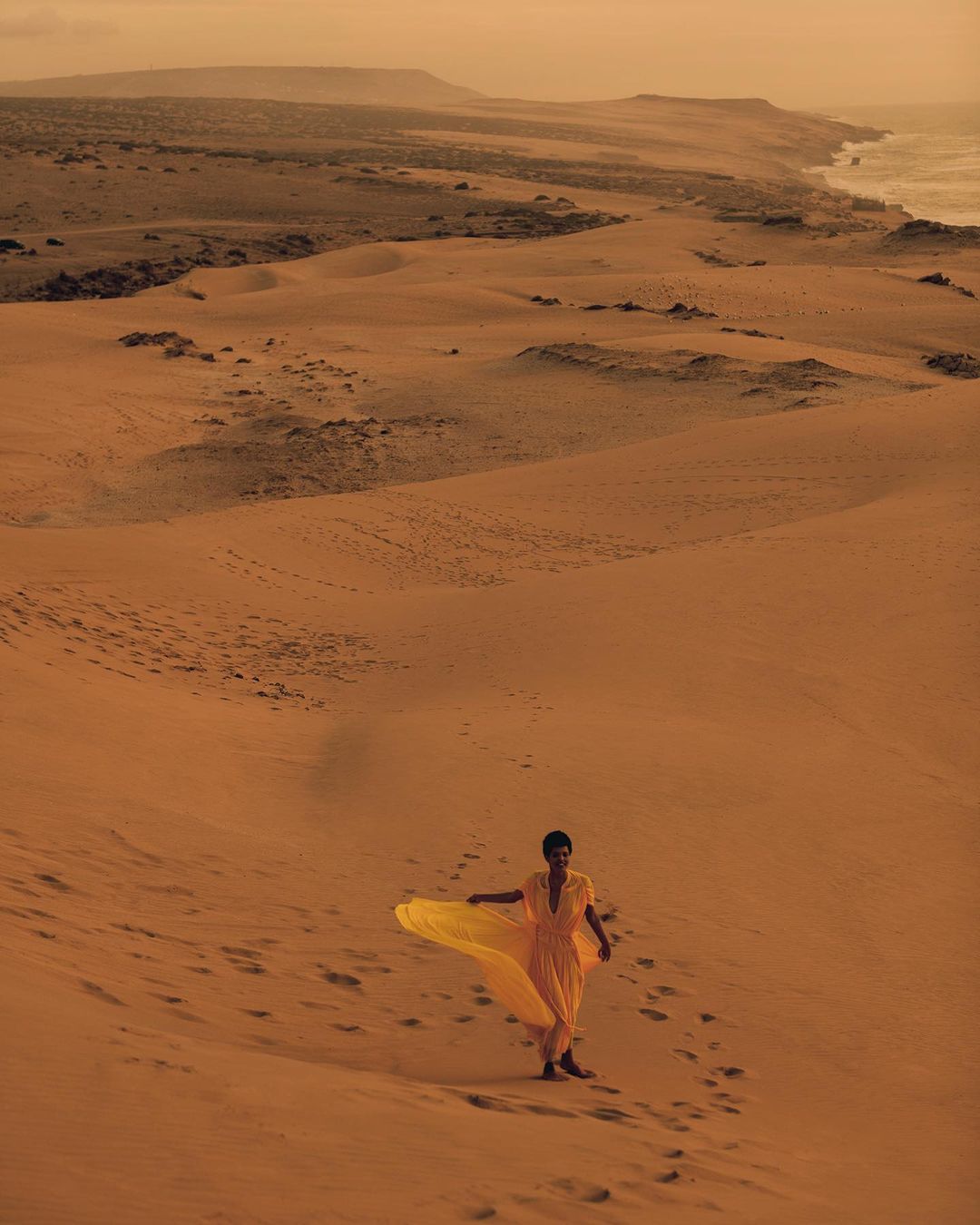 Woman in yellow dress walks on sand dune landscape by the coast, bathed in warm, sandy-hued sunlight, creating a serene desert scene.