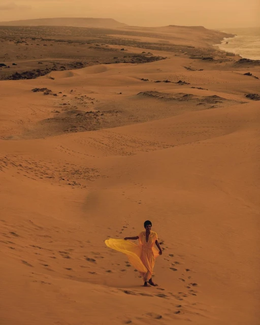 Woman in yellow dress walks on sand dune landscape by the coast, bathed in warm, sandy-hued sunlight, creating a serene desert scene.