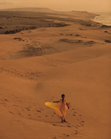 Woman in yellow dress walks on sand dune landscape by the coast, bathed in warm, sandy-hued sunlight, creating a serene desert scene.