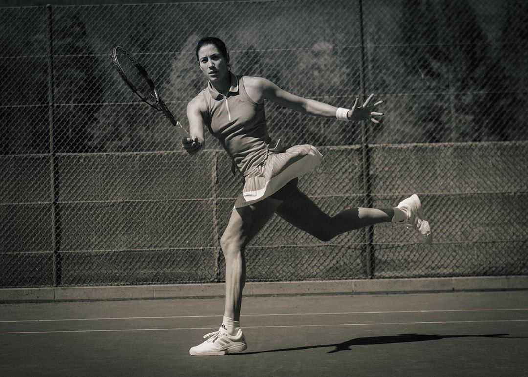 Action shot of a female tennis player in motion on a court, swinging her racquet, with a chain-link fence and trees in the background.