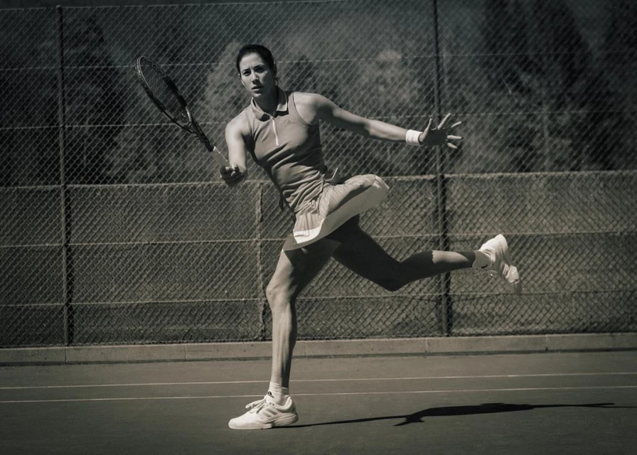 Action shot of a female tennis player in motion on a court, swinging her racquet, with a chain-link fence and trees in the background.