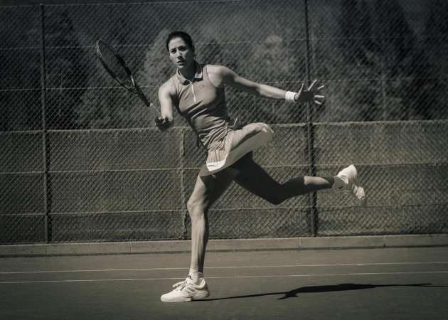 Action shot of a female tennis player in motion on a court, swinging her racquet, with a chain-link fence and trees in the background.