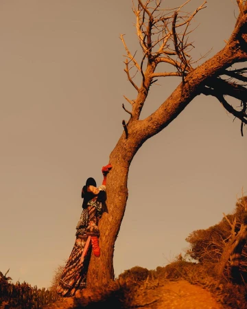A woman in a colorful dress leans against a bare tree in a warm, outdoor setting with a clear sky and earthy tones.