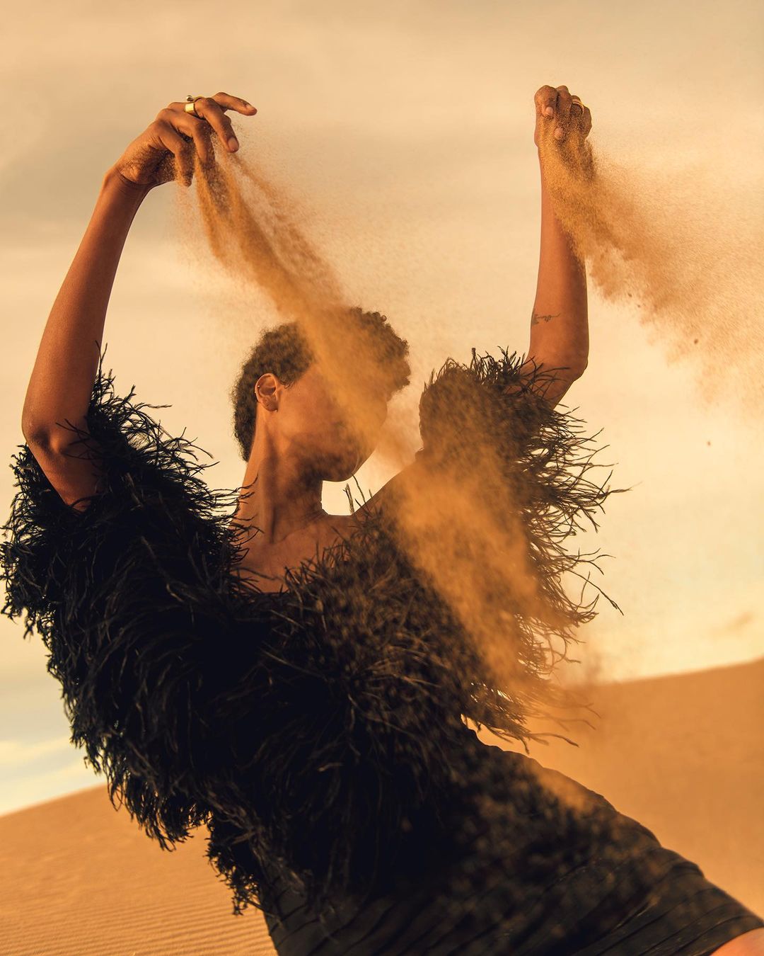 Fashionable woman in a black feathered garment stands in a desert landscape, tossing sand into the air against a cloudy sky.