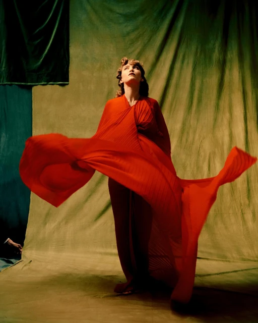 Woman posing in red dress against draped background, studio lighting. Fashion portrait showcases elegant gown, looking upward. Warm, tonal color palette.