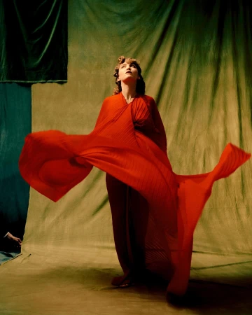 Woman posing in red dress against draped background, studio lighting. Fashion portrait showcases elegant gown, looking upward. Warm, tonal color palette.