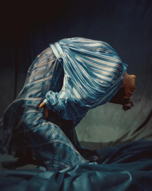 Person in striped blue cloth arches backward in studio, with dramatic lighting and shadows creating a theatrical fashion pose.