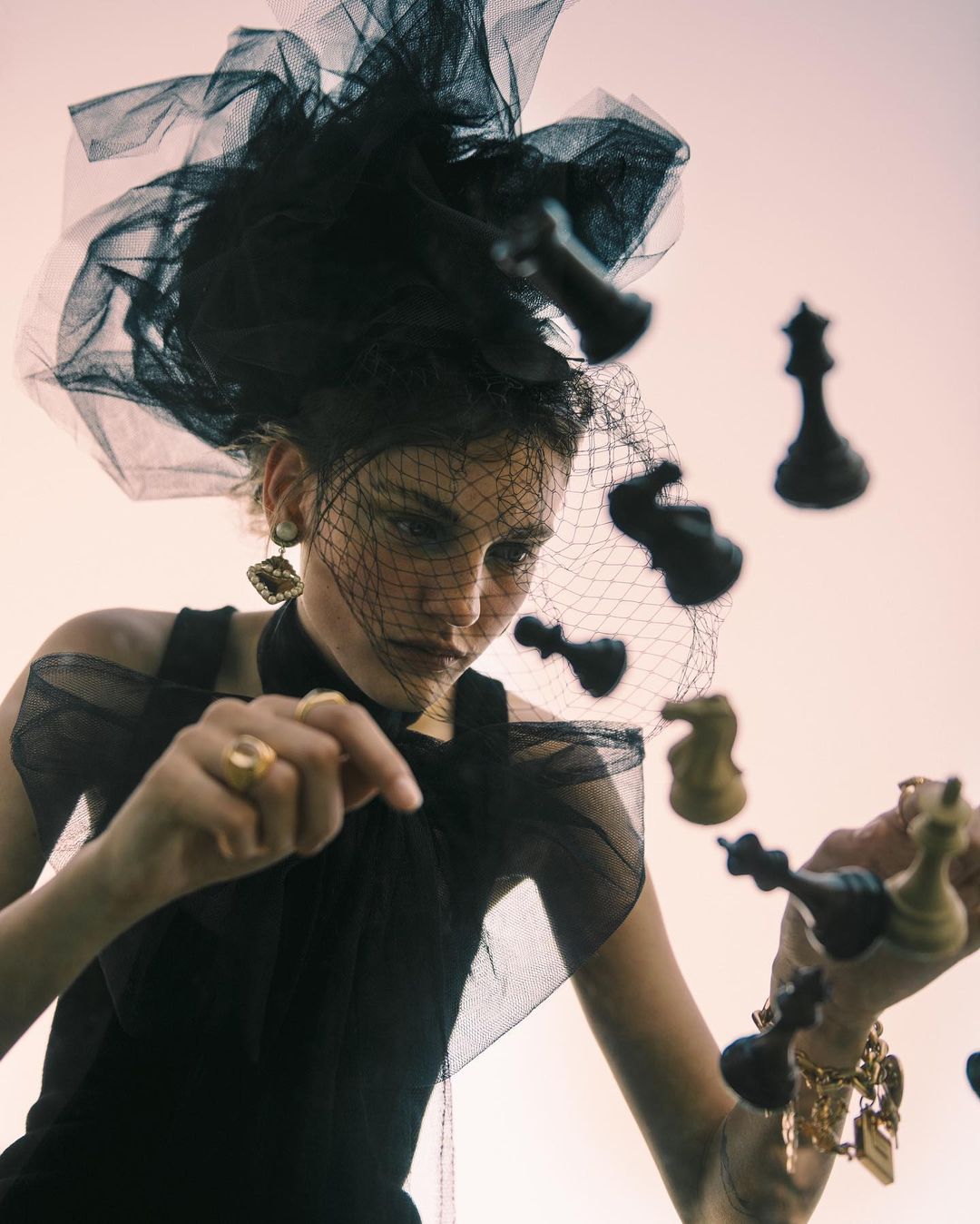 Woman in dramatic black tulle headpiece and dress surrounded by chess pieces in a fashion editorial, shot from a low angle.