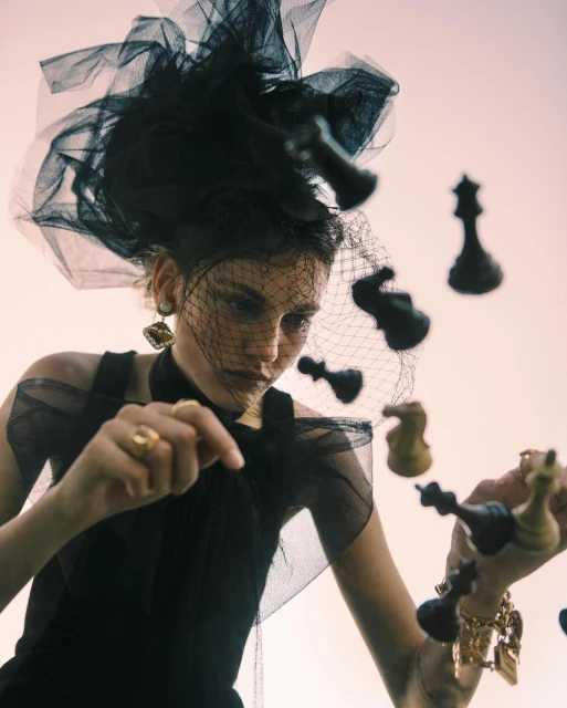 Woman in dramatic black tulle headpiece and dress surrounded by chess pieces in a fashion editorial, shot from a low angle.