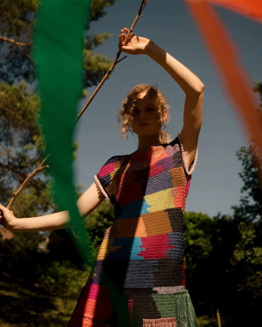 A woman in a colorful patterned dress poses outdoors with a stick against a backdrop of greenery and obscured paint strokes.