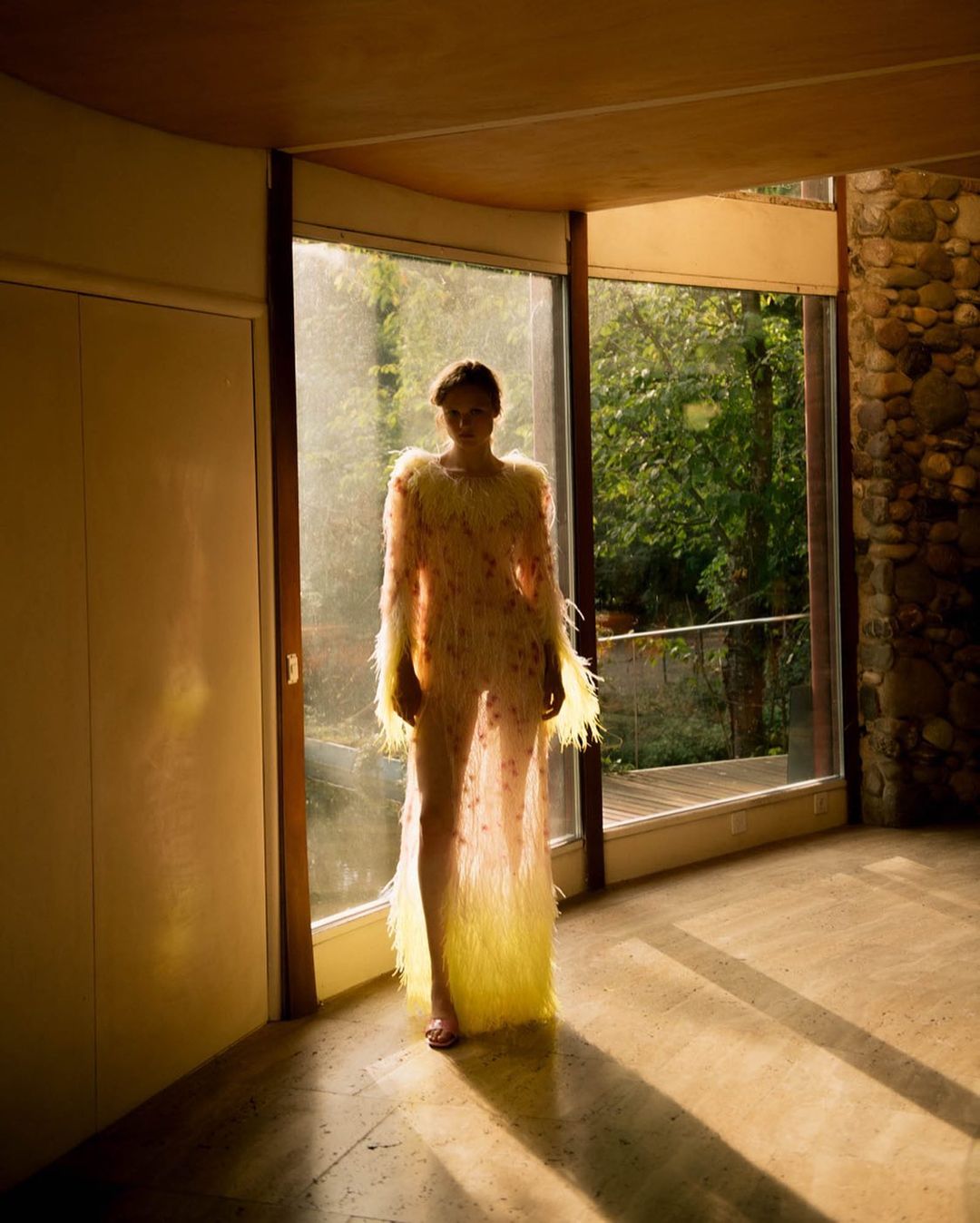 A model poses in a long sheer dress with feather details inside a modern architectural home with stone accents and a large window.