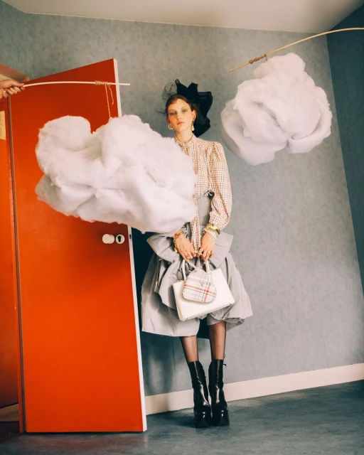 Fashion photo of a woman in a dress and hat holding a purse, with artificial clouds and orange door in a staged indoor setting.