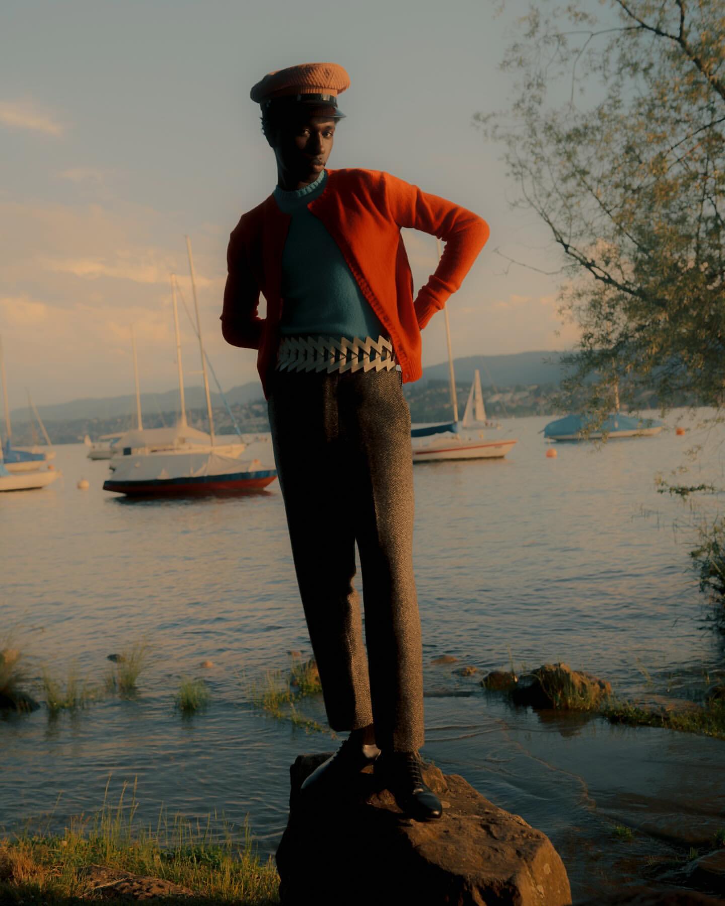 A stylish Black man stands on a rock near a lake with sailboats, wearing an orange cardigan, blue shirt, grey pants, and a tan cap.