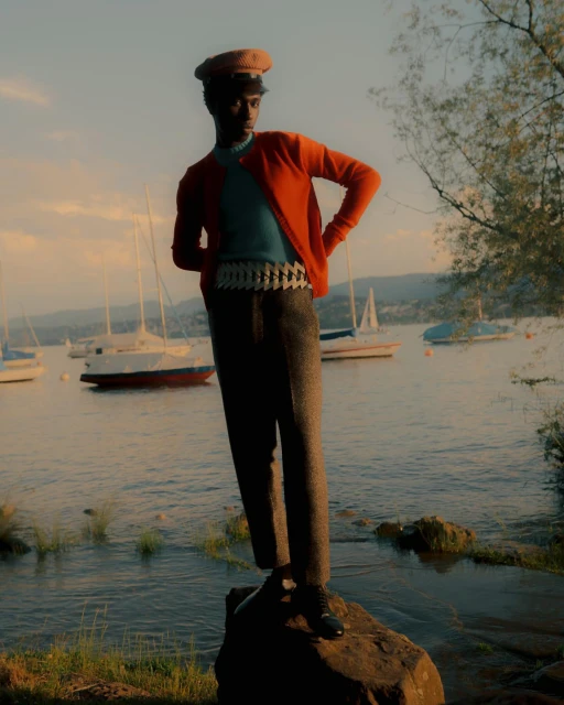 A stylish Black man stands on a rock near a lake with sailboats, wearing an orange cardigan, blue shirt, grey pants, and a tan cap.