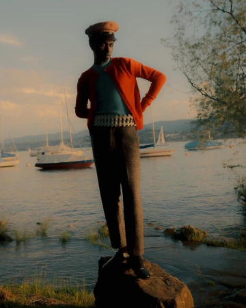 A stylish Black man stands on a rock near a lake with sailboats, wearing an orange cardigan, blue shirt, grey pants, and a tan cap.