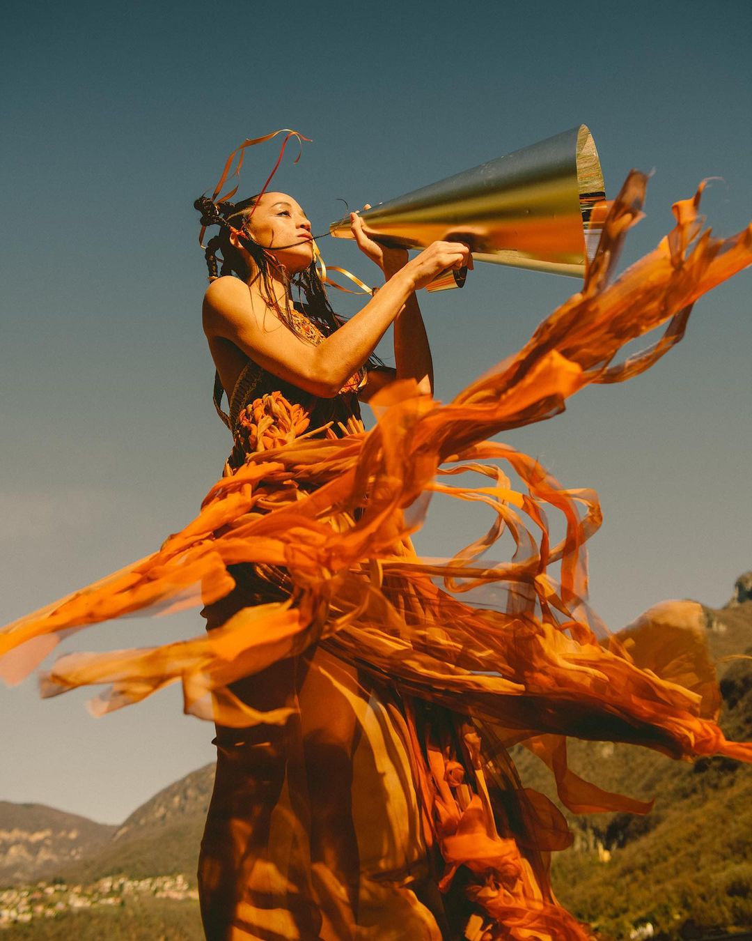 Woman in flowing orange dress speaks into gold megaphone against blue sky and hilly landscape in editorial fashion shot.