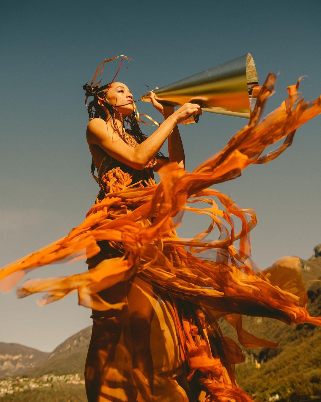 Woman with braided hair outdoors using a gold megaphone and wearing an orange dress with flowing fabric, against a mountain backdrop.