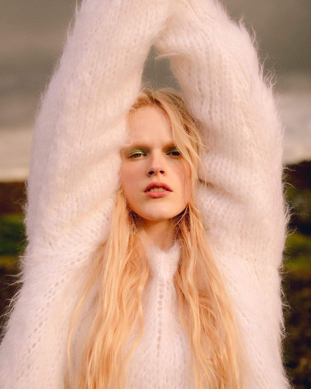 A young adult with long blonde hair poses outdoors wearing a fuzzy white sweater, creating a soft and ethereal fashion portrait.