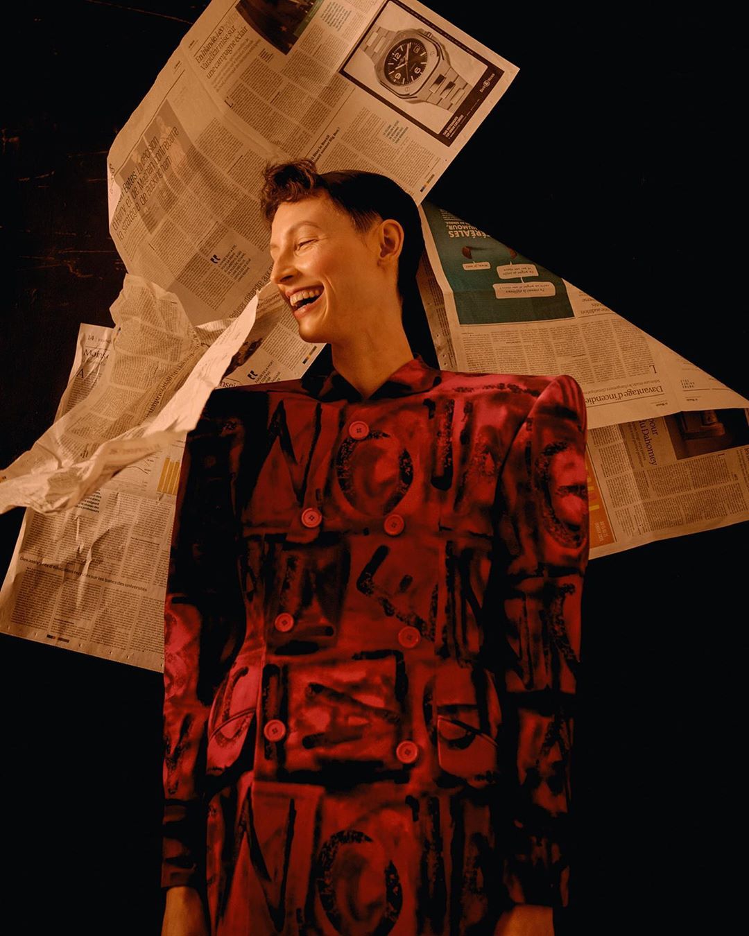 Laughing woman in a red patterned jacket surrounded by newspapers. Portrait shot. Dark background, editorial style photography.