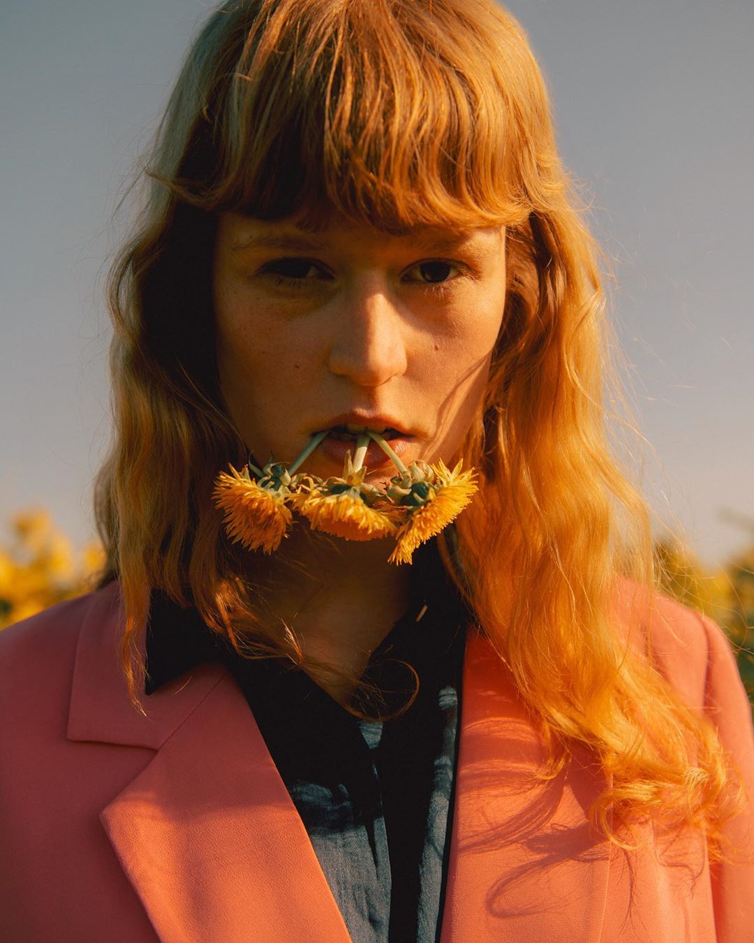 Young woman with red hair and a pink jacket stands outdoors with dandelions in her mouth in a sunlit field, posing for a portrait.