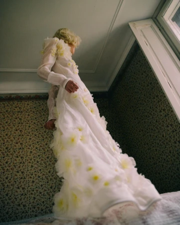 Model in white long-sleeved dress adorned with yellow floral appliqués standing near a patterned wall.