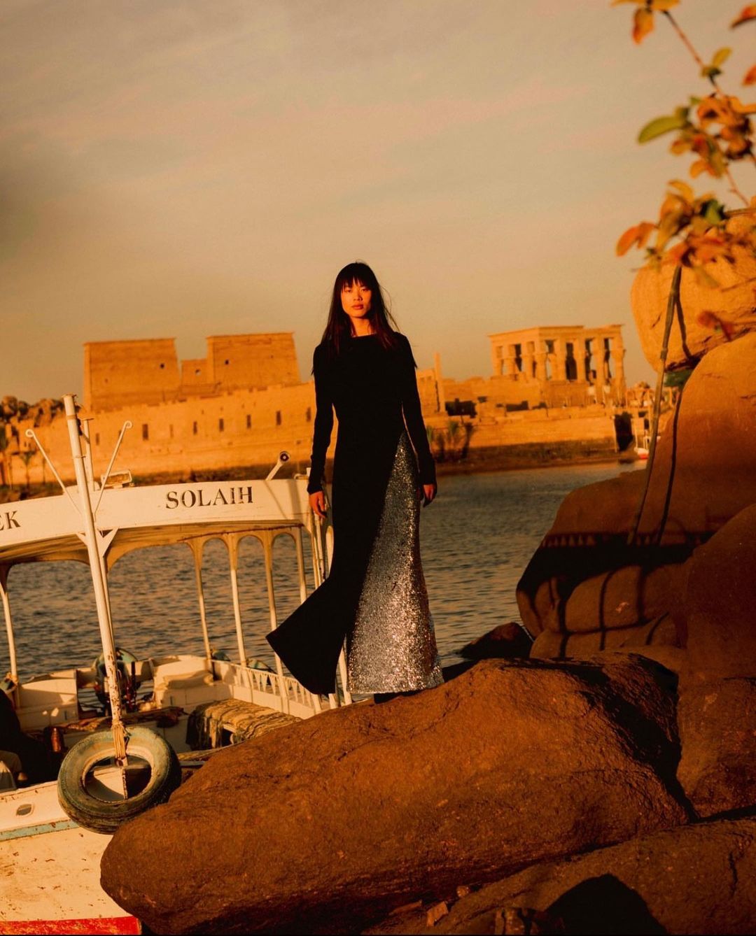 A fashionable woman in a long gown stands on rocky terrain near a river, with an Egyptian temple and a boat visible in the background.